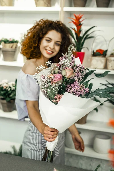 Curly florist with bright lips looking at beautiful flowers in floral shop — Stock Photo, Image