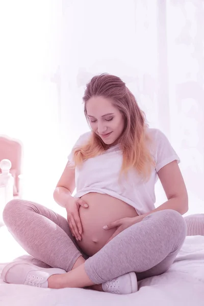 Yatağı üzerinde oturan beyaz tişört uzun saçlı hamile kadında genç — Stok fotoğraf