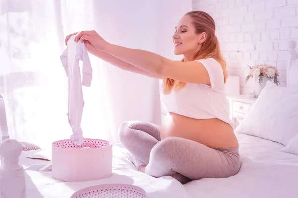 Young long-haired pregnant woman in a white t-shirt showing clothes for a baby — Stock Photo, Image