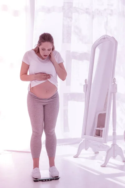 Young beaming long-haired pregnant woman in a white t-shirt standing on the weighing-machine — Stock Photo, Image