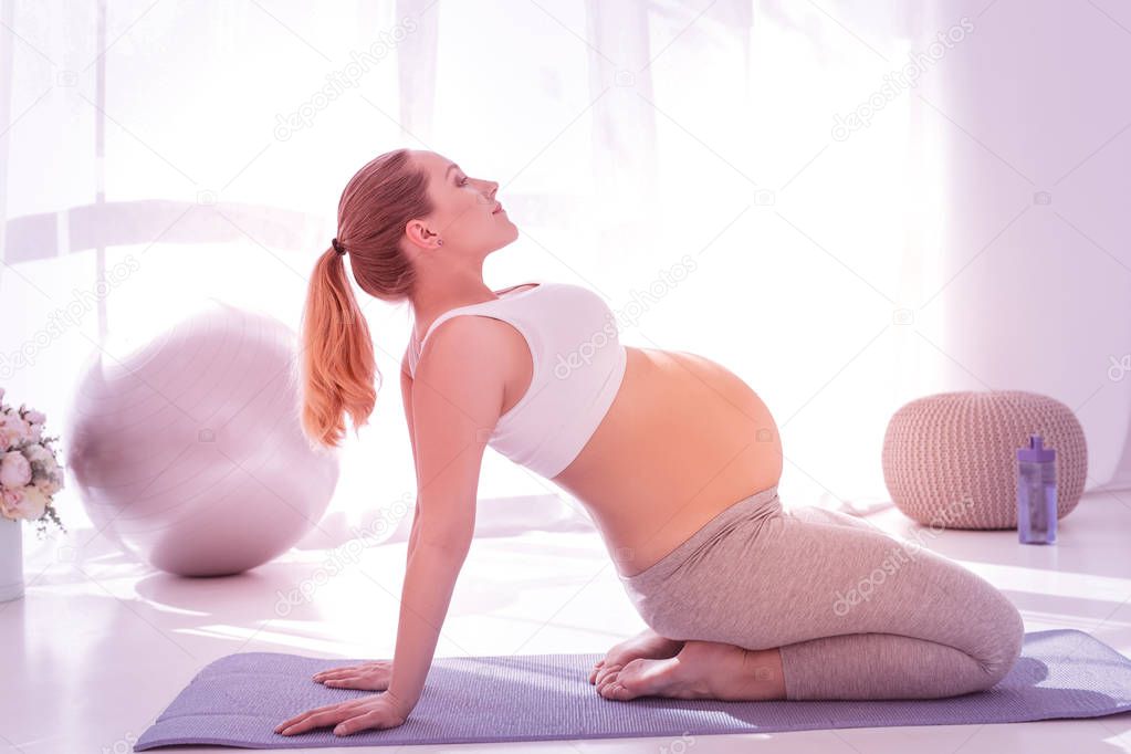 Young beaming long-haired pregnant woman in a white t-shirt practicing yoga