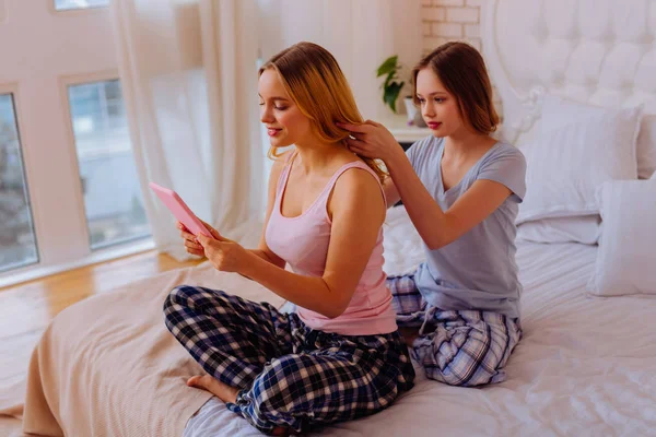 Irmã mais nova fazendo penteado para sua irmã mais velha ocupada usando tablet — Fotografia de Stock