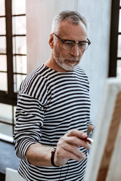 Бородатий художник в окулярах стоїть біля полотна і живопису — стокове фото