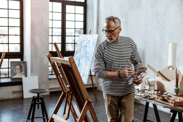 Grey-haired artist looking at painting easel and painting portrait — Stock Photo, Image
