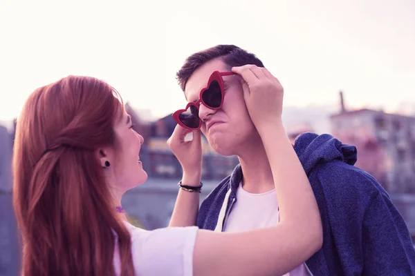 Chica pelirroja sonriente adjuntando gafas de sol rojas funky a la cara de su novio —  Fotos de Stock