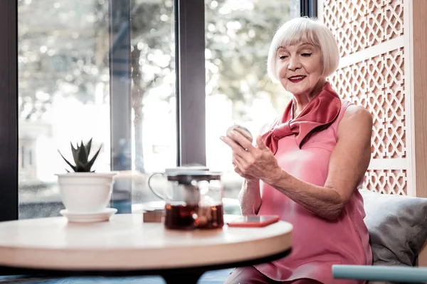 Beaming stylish old lady in pink top checking her makeup — Stock Photo, Image