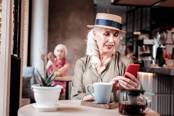 Appealing long-haired old woman in straw hat observing her smartphone