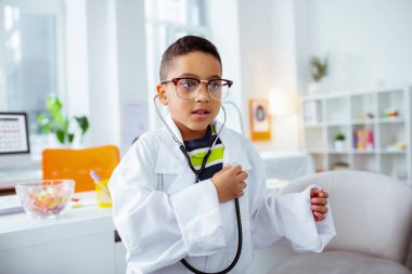 Beyaz ceket ve stetofon giyen çocuk kalbini dinliyor