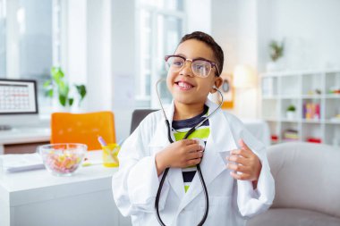 Neşeli çocuk stetofon kullanarak ve doktor beyaz ceket giyiyor