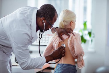 Kız muayene ederken stetoskop kullanarak koyu tenli çocuk doktoru