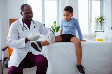 Koyu tenli çocuk doktoru küçük çocuğun bacağına bandaj sürüyor