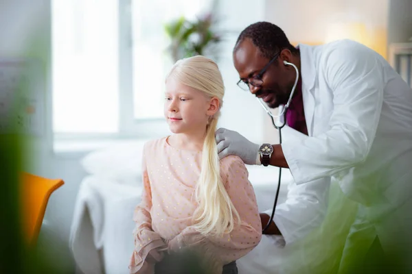 Dark-skinned doctor listening to lungs of girl using stethoscope