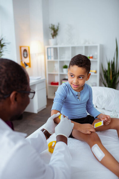 Приятный мальчик, разговаривающий с доктором, намазывающим его тело пластырями
