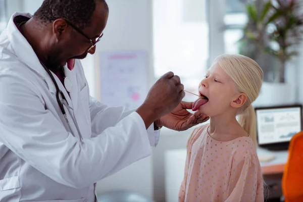 Dark-skinned pediatrician asking little girl to open mouth wide — Stock Photo, Image