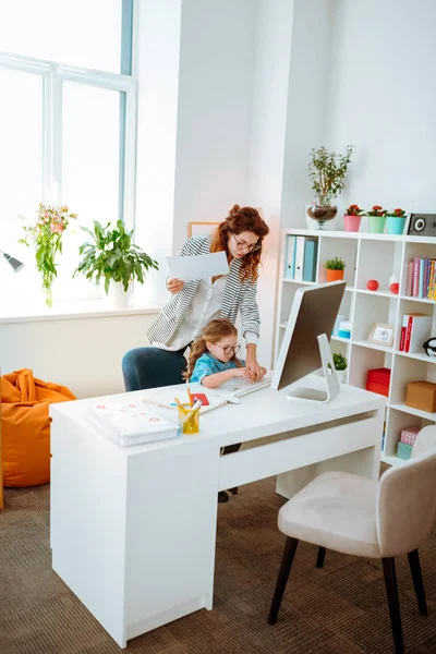 Chica divertida sentada en la mesa de trabajo de su mamá trabajando remotamente — Foto de Stock