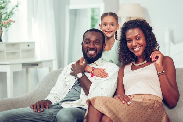 Niña abrazando a padres afro-americanos alegres sentados en el sofá —  Fotos de Stock