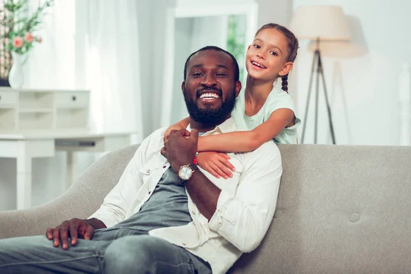 Menina feliz abraçando bonito rindo afro-americano pai no sofá . — Fotografia de Stock