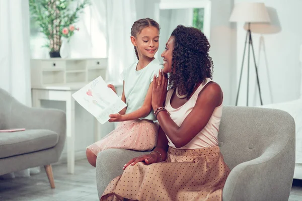 Little Afro-American daughter showing her drawing to beaming cheerful mother — Stock Photo, Image
