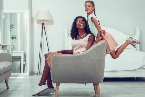 Glowing mother sitting on the chair while daughter having fun. — Stock Photo, Image
