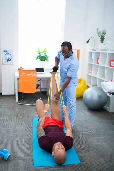 Esportista esticando as pernas no tapete esportivo enquanto visita o terapeuta — Fotografia de Stock