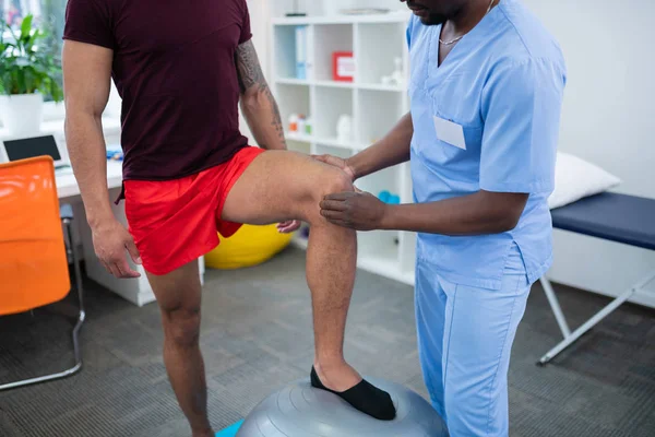 Sportler in roten Shorts legt Bein auf Tannenball, der neben Therapeut steht — Stockfoto