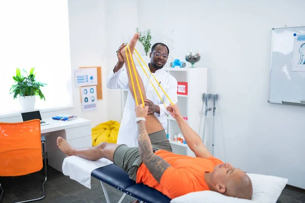 Homem segurando fita amarela ao esticar a perna visitando terapeuta — Fotografia de Stock