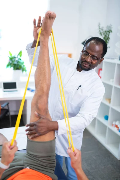 Terapeuta de piel oscura elevando la pierna mientras asiste a deportista — Foto de Stock