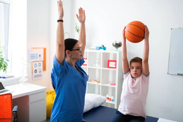 Chateado menino trabalhador sentado no armário médico e levantando bola de basquete — Fotografia de Stock