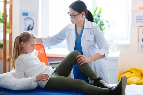 Blondes Kind liegt auf einem Liegebett, während Arzt ihr verletztes Bein beobachtet — Stockfoto