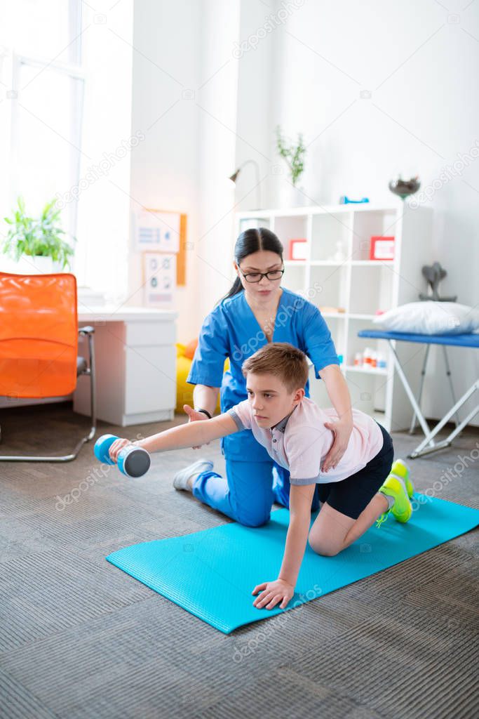 Tranquil short-haired patient holding blue dumbbell while sitting on the mat