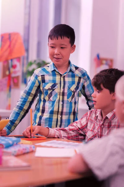 Усміхнений хлопчик стоїть за столом поруч зі своїми однокласниками . — стокове фото