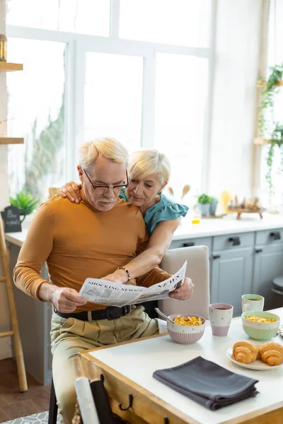 Amar a la esposa tiernamente abrazar a un cónyuge durante la lectura del periódico — Foto de Stock