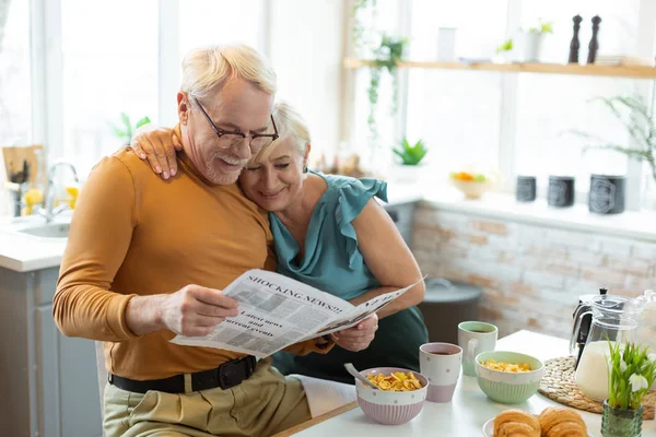 Посміхаючись приємно приваблива у віці біло-волохата подружня пара читає газету разом — стокове фото
