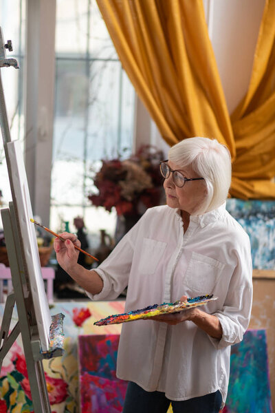 Blonde-haired retired artist standing near window and painting