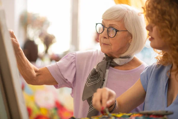 Aged artist helping her curly student with picture — Stock Photo, Image