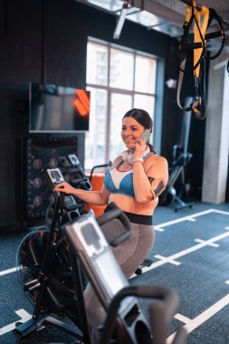 Spor salonunda bisiklet ve telefonda konuşan koyu saçlı kadın