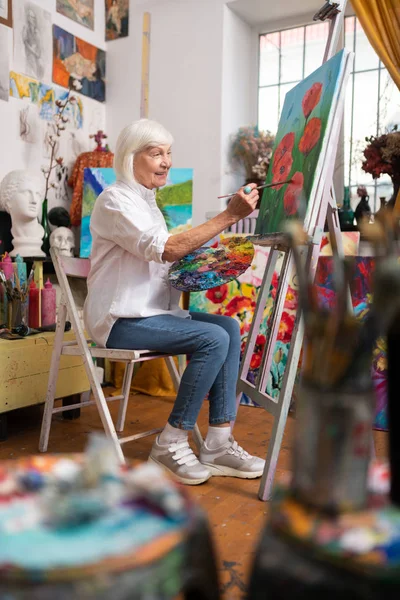 Mulher idosa vestindo jeans sentado e pintando sobre tela — Fotografia de Stock