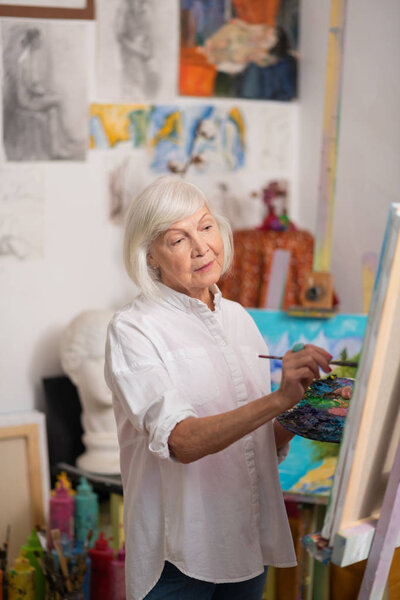 Пожилая женщина стоит возле рисования мольберта и наслаждается своим хобби
