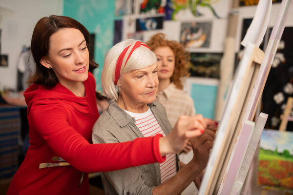 Aged art teacher and her students helping her drawing on canvas