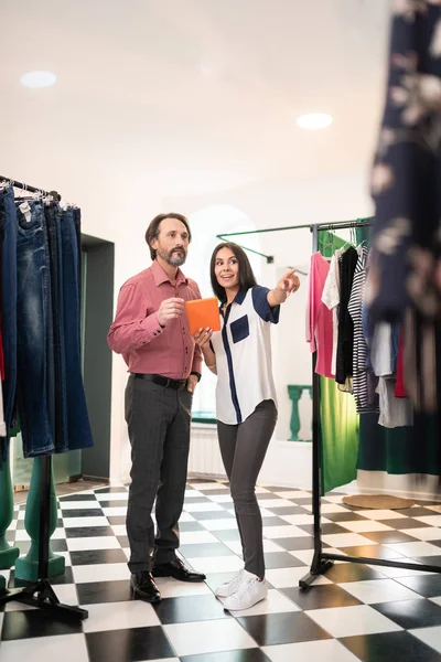 Cheerful stylist pointing at clothing while working with focused customer.