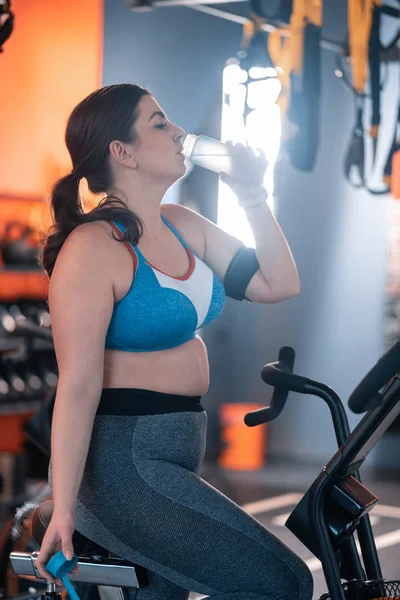 Mujer con sobrepeso bebiendo agua mientras pedalea en el gimnasio — Foto de Stock