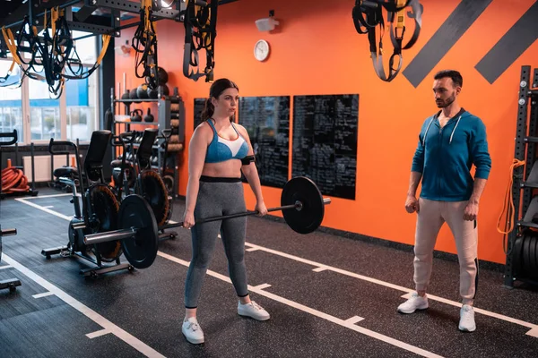 Frau hält Langhantel, während sie mit Trainer im Fitnessstudio trainiert — Stockfoto