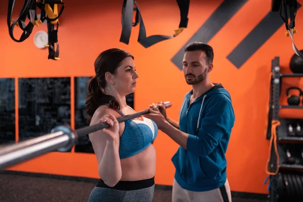 Bearded trainer assisting plump woman with lifting barbell