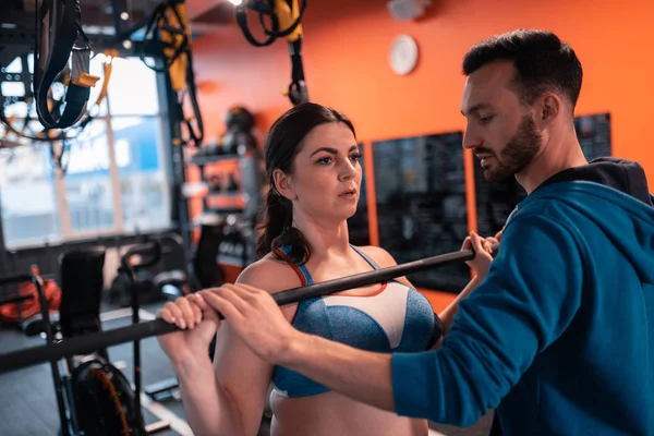 Mujer morena haciendo ejercicio con la barra cerca de su entrenador — Foto de Stock