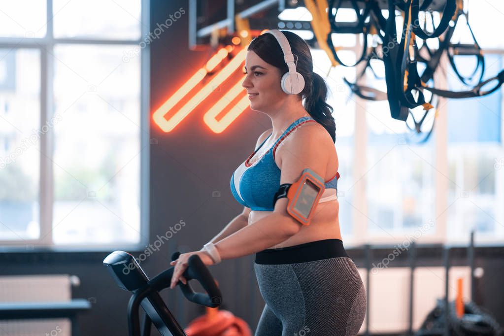 Overweight woman cycling in gym and listening to music in earphones