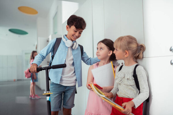 Dark-haired boy standing near scooter speaking with classmates