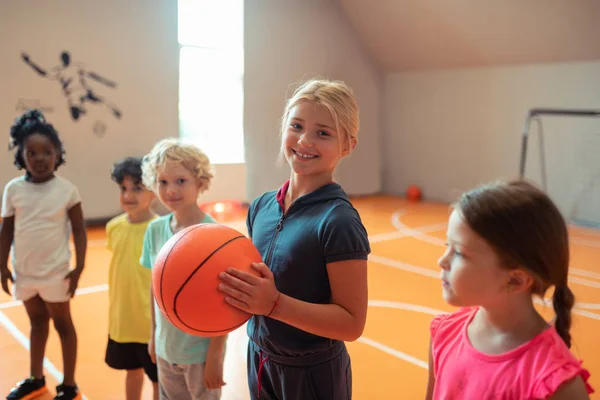 Sorrindo menina segurando uma bola de pé ao lado de seus colegas de classe . — Fotografia de Stock