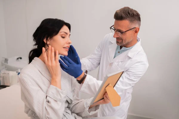 Schönheitschirurg mit Brille berührt Gesicht des Patienten nach Operation — Stockfoto