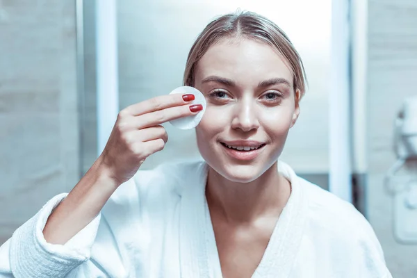 Good-looking woman with soft smile cleaning face with soaked cotton pad