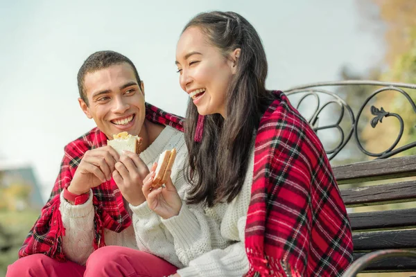 Glädjefullt snygg par som äter nybakade smörgåsar på en bänk — Stockfoto